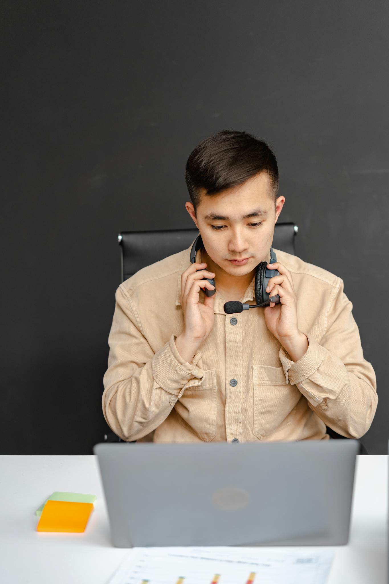 Serious Asian man in call center office, wearing a headset and working on a laptop.